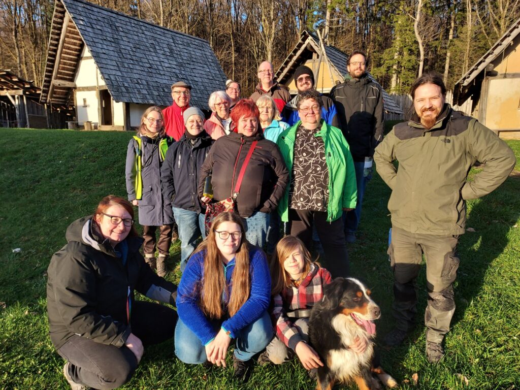 Eine Gruppe von 16 Personen steht im Freien, umgeben von Bäumen und historischen Gebäuden, mit einem Hund im Vordergrund.