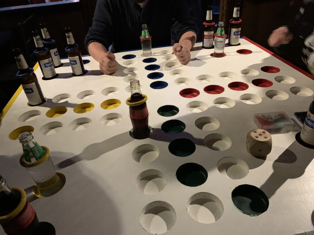 Ein Tisch mit einem Brettspiel, Flaschen und einem Würfel, das von mehreren Personen genutzt wird.