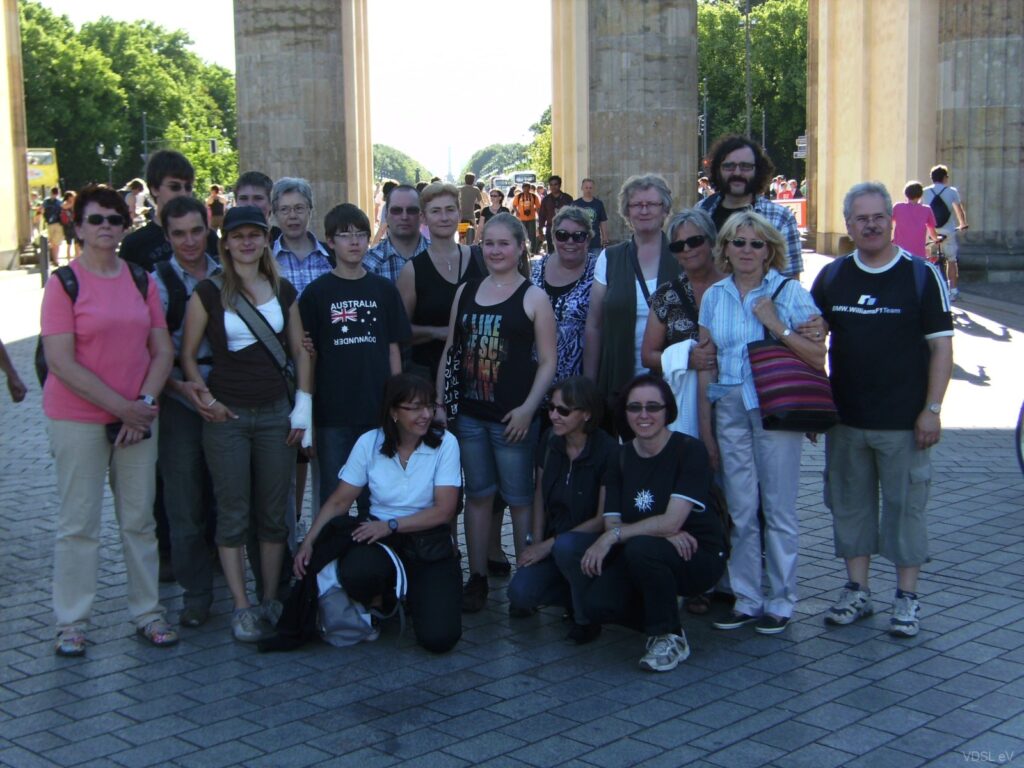 Eine Gruppe von 20 Personen posiert vor dem Brandenburger Tor in Berlin an einem sonnigen Tag.