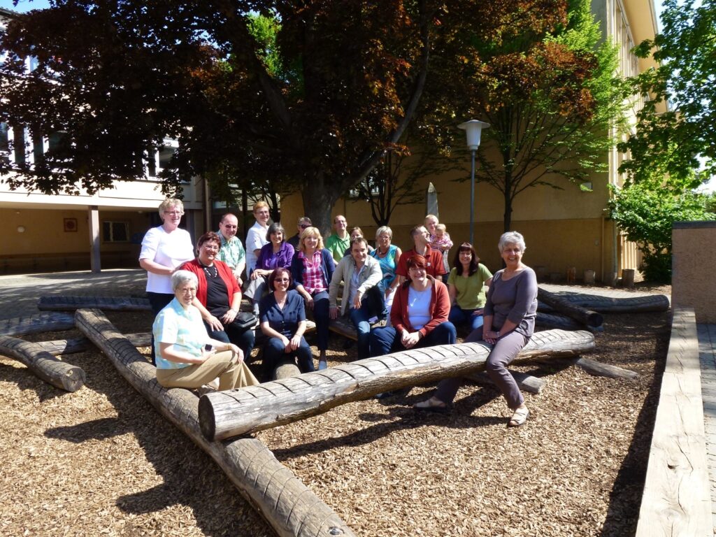 Eine Gruppe von 16 Personen sitzt auf Holzstämmen in einem Außenbereich mit Bäumen.