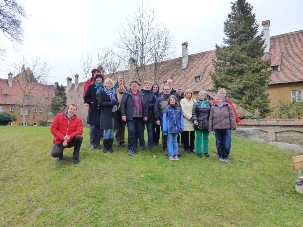 Eine Gruppe von Personen steht auf einer Wiese vor einem Gebäude mit roten Ziegeldächern.
