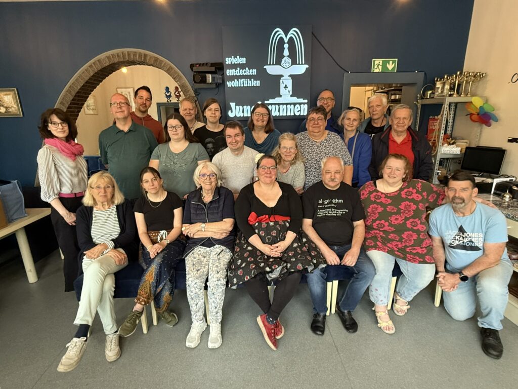 Gruppe von Teilnehmenden beim Symposium 2025 in Nettetal im Dorf- und Spielecafé Jungbrunnen; Petra und Erwin sind sichtbar vor einer dunkelblauen Wand mit Logo