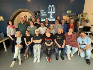 Gruppe von Teilnehmenden beim Symposium 2025 in Nettetal im Dorf- und Spielecafé Jungbrunnen; Petra und Erwin sind sichtbar vor einer dunkelblauen Wand mit Logo
